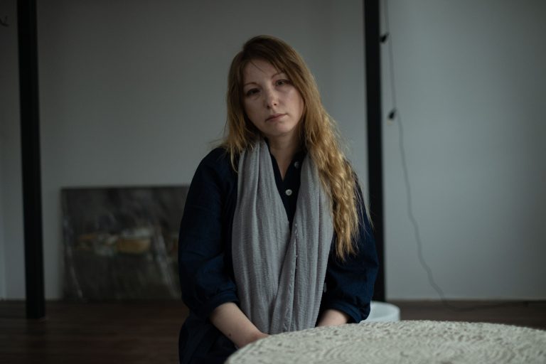 A woman with a gray scarf sits at a table in a gallery, looking at the camera.