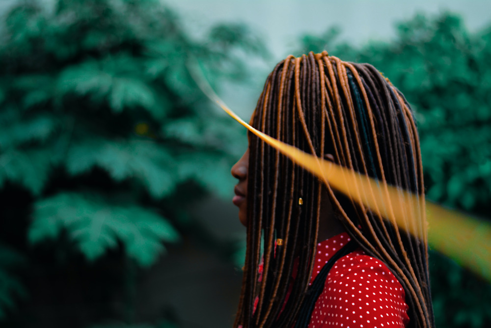 A woman with dreadlocks and her face obscured stands outside, surrounded by trees